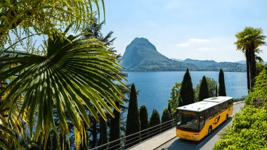 A yellow bus is driving on a road alongside a lake, surrounded by lush palm trees and mountains in the background.