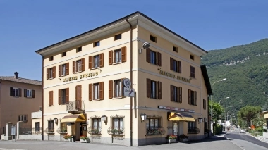 Un bâtiment jaune à trois étages avec des volets et l'inscription "Albergo Svizzero", entouré de montagnes.