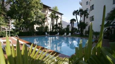 Piscina all'aperto con lettini, circondata da palme ed edifici bianchi, montagne sullo sfondo.