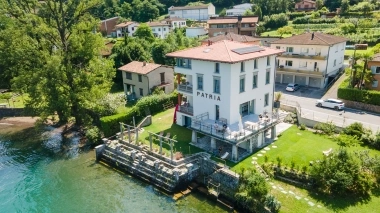 Aerial view of a large building with the sign "PATRIA" by the water, surrounded by greenery and residential houses.