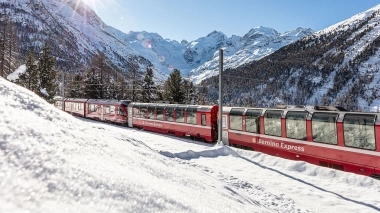 A red Bernina Express train travels through a snowy mountain landscape in sunshine.