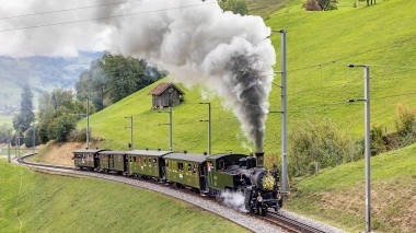 Eine dampfende Dampflokomotive zieht grüne Waggons durch eine hügelige Landschaft.