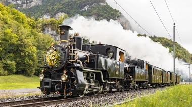 A steaming historic locomotive adorned with flowers travels through a green landscape.