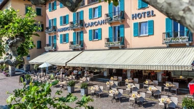 Hotel con persiane turchesi, terrazza del caffè e tende, circondato da alberi.