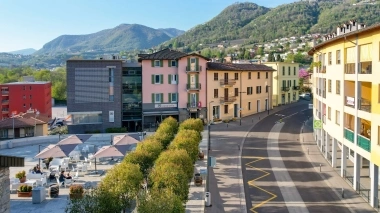 A pretty street with buildings, cafés with umbrellas, and mountains in the background.