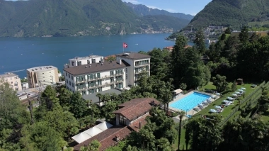 Luftaufnahme eines Hotels mit Pool, umgeben von Bäumen, vor einem grossen See und bewaldeten Bergen.