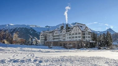 A large white hotel building in the snow in front of a mountain backdrop under a blue sky.