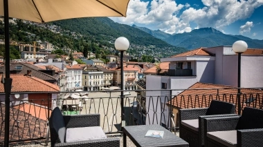 Eine Terrasse mit Blick auf die Dächer einer Stadt, umgeben von Bergen unter blauem Himmel.