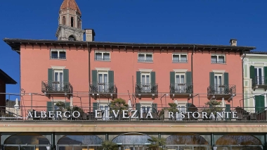 Eine rosafarbene Gebäudefassade mit mehreren Balkonen und der Aufschrift "Albergo Elvezia Ristorante" im Vordergrund.