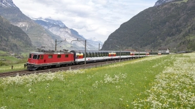 Eine rote Lokomotive zieht einen Zug durch eine grüne Berglandschaft.