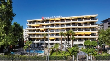 Multi-story hotel building with balconies, pool, and palm trees in the foreground.