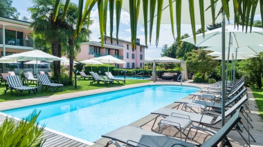 Zone de piscine ensoleillée avec des chaises longues, des parasols et des palmiers à côté d'un bâtiment rose.