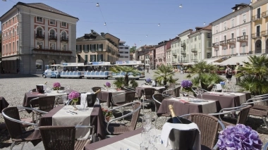 A pretty piazza with outdoor tables, flowers, and a tourist train passing by colorful house fronts.