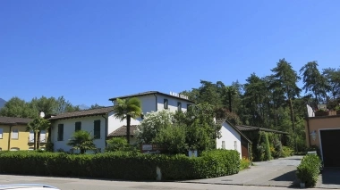 A white building with green shutters, surrounded by trees and plants, under a clear blue sky.