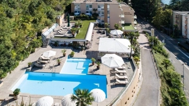 Aerial view of a hotel with a pool, sun umbrellas, and sun loungers, surrounded by green hills.