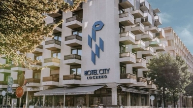 Bâtiment à plusieurs étages avec balcons, enseigne "Hotel City Locarno", entouré d'arbres.