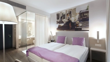 Modern hotel room with double bed, purple cushions and blanket, glass-walled bathroom, and large wall mural.