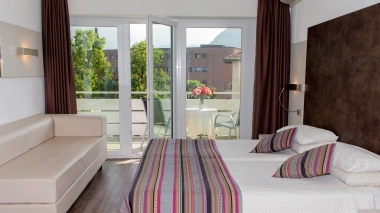 A hotel room with a double bed, sofa, balcony with table and flowers, and large windows overlooking trees and buildings.