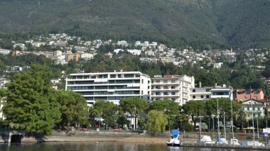 Eine Stadt am Hang eines grünen Hügels mit modernen Gebäuden und Segelbooten am Wasser.