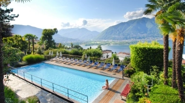 A woman sits at the edge of a pool overlooking a lake and surrounding mountains, surrounded by palm trees and green vegetation.