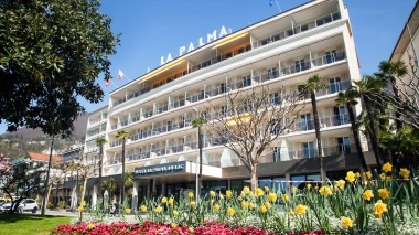 A multi-story hotel building with balconies, surrounded by colorful flower beds and palm trees, under a clear sky.