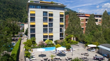 Bâtiment à plusieurs étages avec des auvents jaunes, piscine et parasols au premier plan, entouré d'arbres et de montagnes à l'arrière-plan.