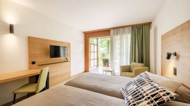 Bright hotel room with two beds, a desk, a TV, and a balcony with a view of the greenery.