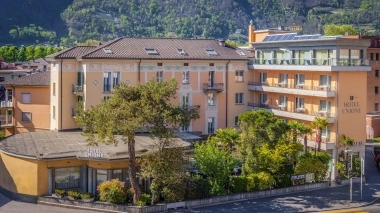 Un edificio alberghiero a più piani con facciata gialla, circondato da alberi verdi e montagne sullo sfondo.