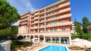 A multi-story pink building with balconies, a pool in the foreground, and lounge chairs under umbrellas.