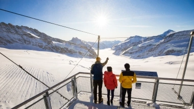 A group of three people on a viewing platform overlooking a snowy mountain landscape.