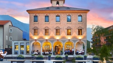 Historisches Gebäude mit beleuchtetem Restaurant im Erdgeschoss, umgeben von Sitzgelegenheiten im Freien bei Sonnenuntergang.