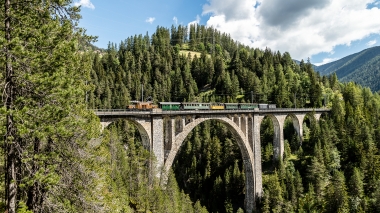 Eine historische Eisenbahn fährt über ein steinernes Viadukt inmitten eines bewaldeten Tals.