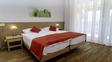 Une chambre d'hôtel avec un grand lit double, des coussins et un plaid rouges, deux tables de chevet avec des lampes et un tableau de pommes vertes au mur.