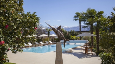 Espace piscine ensoleillé avec transats, palmiers et une sculpture d'espadon devant un décor montagneux.