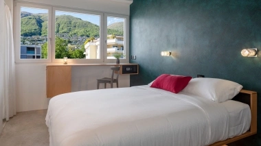 A modern bedroom with a white bed, dark green wall, large window, and mountain view.