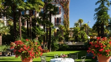 Garten mit Tisch und Stühlen, umgeben von Palmen und einem alten Gebäude im Hintergrund.