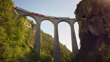 A red panoramic train crosses the impressive Landwasser Viaduct, an iconic structure nestled in a lush green mountain landscape.