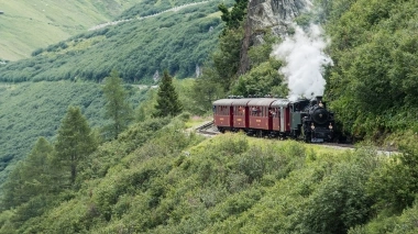 A steaming train travels through a green mountain landscape.