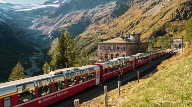 A red panoramic train travels through an alpine landscape with high mountains and a stone house.