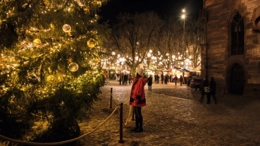A person stands in front of a large, illuminated Christmas tree in a festive square at night.