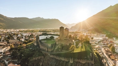 Eine alte Burg auf einem Hügel mit zwei Türmen, umgeben von einer modernen Stadtlandschaft bei Sonnenuntergang.