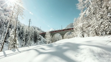 Eine rote Bahn überquert eine steinerne Brücke in einer verschneiten Berglandschaft.