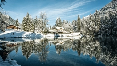 Un paysage hivernal avec une maison au bord d'un lac enneigé, entourée d'arbres recouverts de neige.