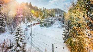 A winter landscape with a train traveling through snowy forests in sunlight.