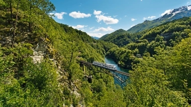 Eine Eisenbahnbrücke überquert eine Schlucht in einer grünen, bergigen Landschaft unter klarem, blauem Himmel.