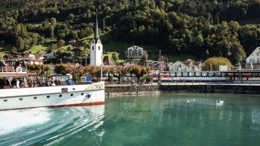 Un ferry sur un lac, avec une église et des maisons dans un paysage de montagne verdoyant en arrière-plan.
