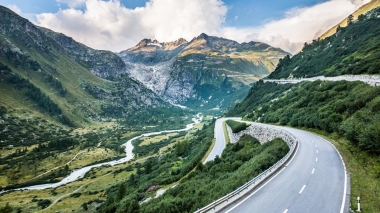 Eine kurvige Bergstrasse, die durch ein grünes Tal in Richtung imposanter Berggipfel führt, mit bewölktem Himmel im Hintergrund.