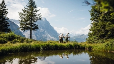 Due persone che camminano su un sentiero di montagna con alberi e un lago in primo piano.