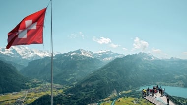 A group of people on a viewing platform overlooking a valley and mountains, next to a Swiss flag.