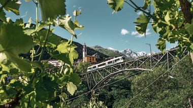 A train is crossing a bridge surrounded by green leaves, with a church steeple and snow-capped mountains in the background.